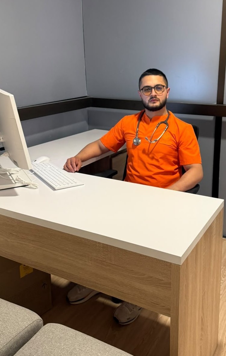 Profesional de la salud de Vital Care con uniforme médico naranja, estetoscopio y lentes, sentado tras su escritorio en el consultorio